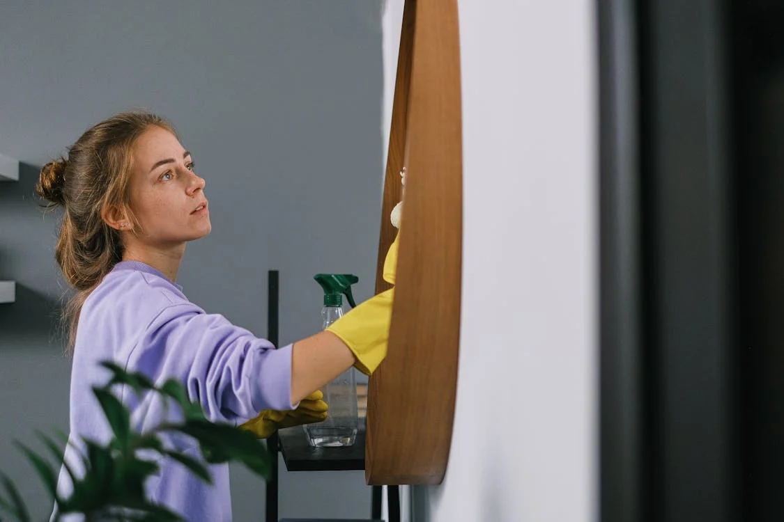 Pensive female cleaning mirror in light room with gray walls